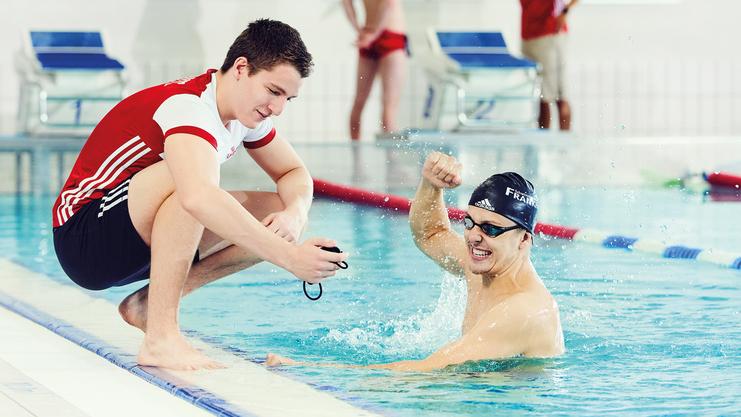 Schwimmtrainer zeigt Stoppuhr, Sportler freut sich über die Zeit.